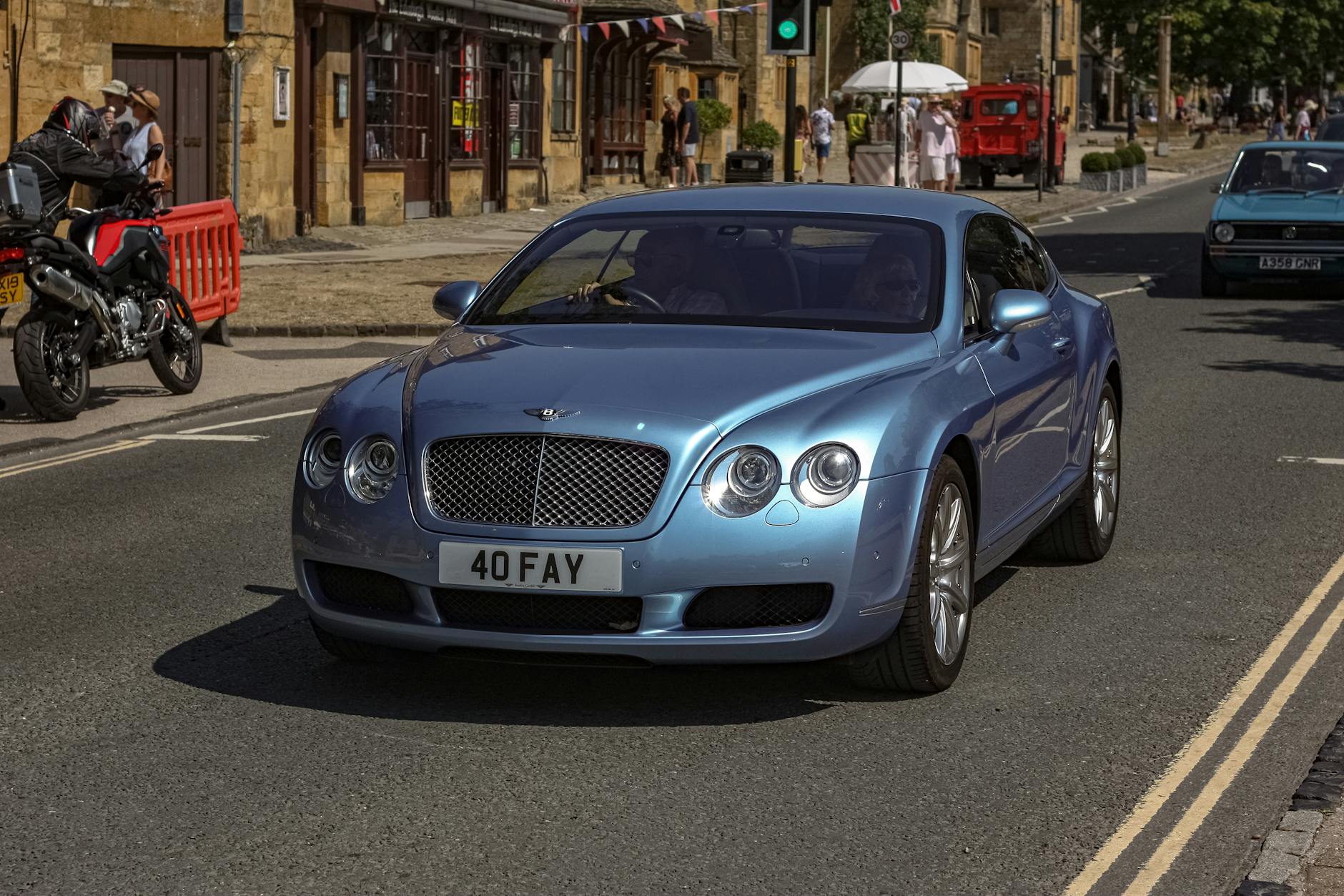 Niebieski Bentley Continental jedzie słoneczną ulicą obok pieszych i motocykli